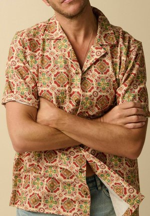 Man met gekruiste armen, gekleed in een korte mouw blouse met een rood, groen en geel geometrisch bloemmotief.