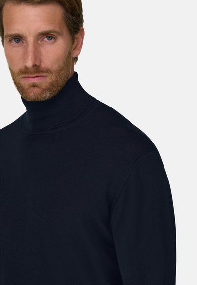 Homme avec une légère barbe portant un pull à col roulé bleu marine foncé, tourné légèrement vers la gauche, expression neutre sur un fond clair uni.