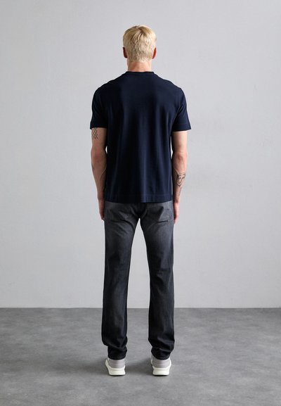 T-shirt navy a maniche corte in cotone, abbinato a jeans dritti grigio scuro e sneakers chiare, visto da dietro.