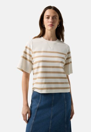 Woman wearing a cream and beige striped short-sleeve top paired with a blue high-waisted denim skirt standing against a plain background.