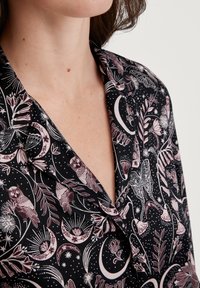 De bovenkant van een vrouwelijk figuur, met een zwart shirt dat is versierd met een patroon van uilen, halve manen, sterren, bloemen en vlinders in wit en gedempt paars.
