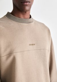Man wearing a beige crewneck sweatshirt with ribbed collar and subtle "BOSS" logo embroidered on chest against a plain background.