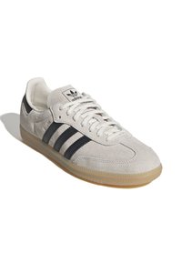 Baskets en daim gris clair avec des rayures en cuir noir, une semelle en caoutchouc couleur gomme, et des lacets blancs. Présente le logo Adidas sur la languette.