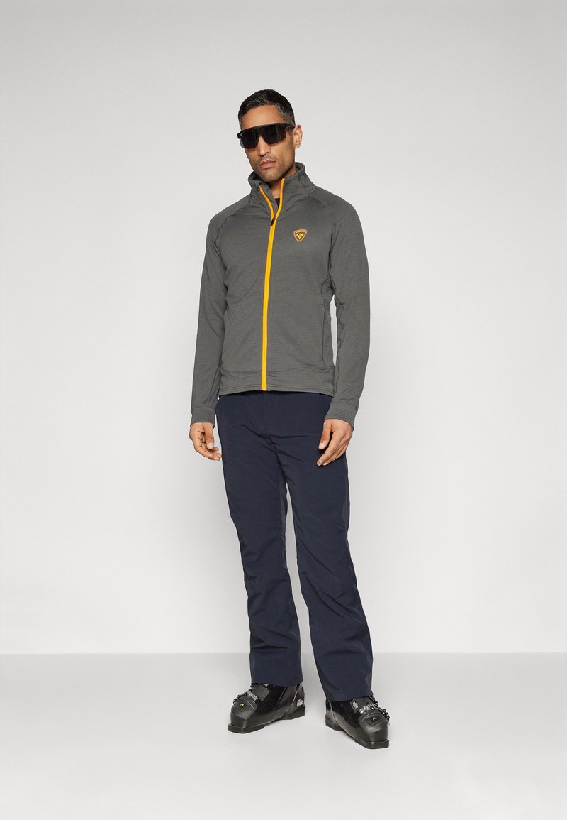 Szara kurtka zapinana na zamek z pomarańczowymi akcentami, teksturowana tkanina i logo na piersi; zestawiona z granatowymi spodniami i czarnym obuwiem.