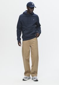 Marineblå hættejakke med forlommer, beige bukser og grå sneakers. Kappen har et hvidt logo. Enkel, afslappet pasform, minimal detaljer.
