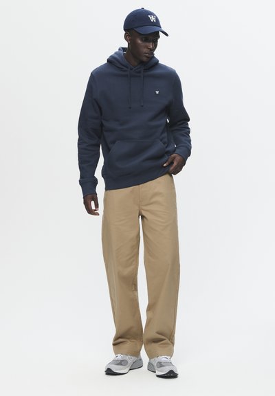 Sweat à capuche bleu marine avec poche avant, pantalon beige et baskets grises. La casquette arbore un logo blanc. Style simple, coupe décontractée, peu de détails.