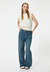 Top sans manches en tricot blanc avec des motifs texturés, associé à un jean en denim bleu ample. Le jean présente un tissage en denim traditionnel et cinq poches.