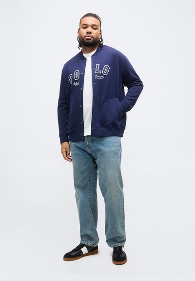 Άνδρας με πλεξούδες που φορά ναυτικό μπλε σακάκι με κουμπιά, λευκή μπλούζα, ανοιχτό μπλε τζιν και μαύρα αθλητικά παπούτσια με άσπρες ρίγες.