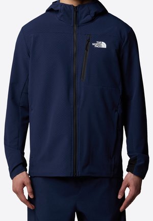 Mand iført en mørkeblå jakke med lynlås og hætte og The North Face-logo på venstre bryst, hænderne afslappet langs siden.