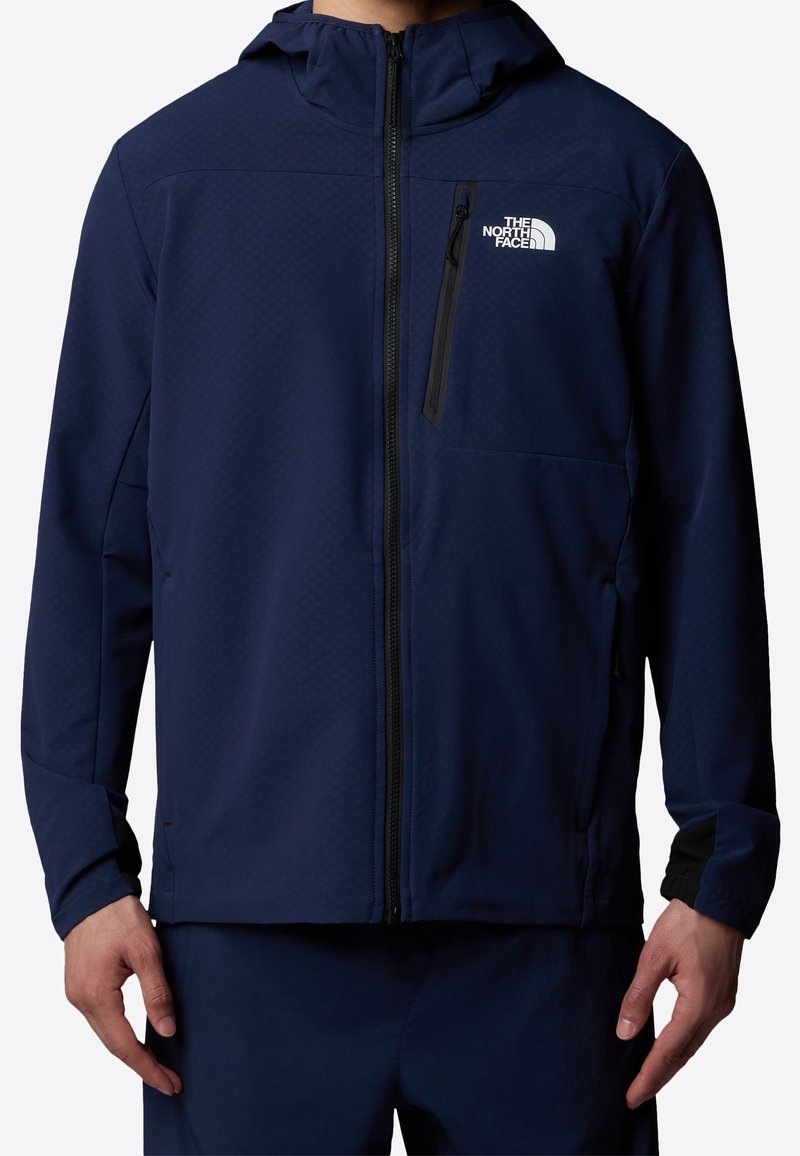 Homme portant une veste zippée bleu marine avec capuche et le logo The North Face sur la poitrine gauche, les mains détendues le long du corps.
