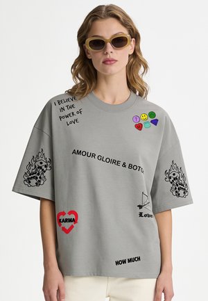 Frau trägt übergroßes graues T-Shirt mit aufgedruckten Phrasen, Emojis, brennenden Würfeln und Herzgrafiken, kombiniert mit beigen Hosen und gelber Sonnenbrille.