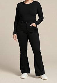 Personne portant un haut noir moulant à manches longues, un pantalon noir évasé taille haute et des baskets blanches, debout avec une main dans la poche.