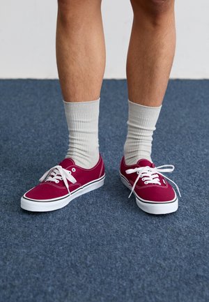 Rote Leinenschuhe mit weißen Schnürsenkeln und schwarzem Rand, kombiniert mit hellgrauen gerippten Socken. Platziert auf einer blauen strukturierten Oberfläche.