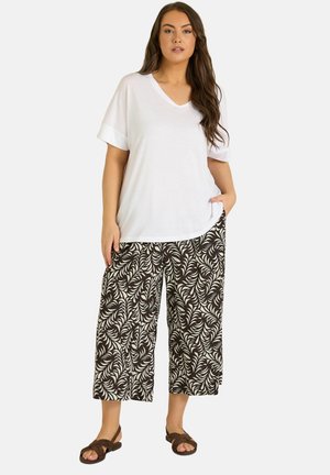 Donna che indossa una maglietta bianca a maniche corte con scollo a V, pantaloni a gamba larga e corta con motivo bianco e nero, e sandali marroni, in piedi con una mano in tasca.