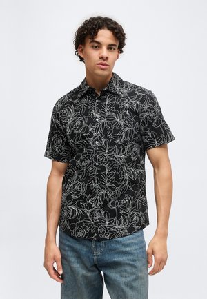 Jeune homme portant une chemise noire à manches courtes avec des contours floraux blancs et un jean bleu, debout devant un fond uni.