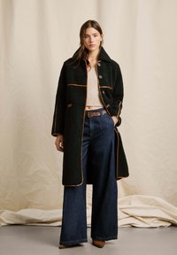 Cappotto verde con texture e piping arancione, dotato di colletto alto e chiusura con bottoni, abbinato a jeans in denim a vita alta e gamba larga.
