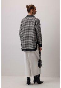 Crna i bijela jakna u uzorku houndstooth s crnim satenastim ovratnikom, nošena preko bijele suknje srednje dužine, u kombinaciji s crnim čizmama s oštrim vrhom.