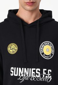 Čierna mikina s kapucňou a šnúrkami. Obsahuje žlté a biele okrúhle nášivky s logom a biely text s nápismi "SUNNIES FC" a "Lyle & Scott."