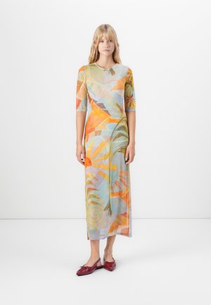 Vrouw die staat in een kleurrijke, lange jurk met lange mouwen tot op de enkels met een abstract bloemmotief, en rode puntige flats draagt.