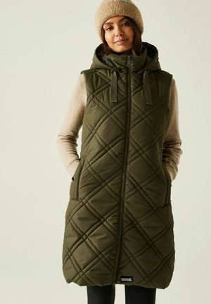 Woman wearing a quilted olive green hooded sleeveless vest over a beige sweater and beige knit beanie, with hands in vest pockets.