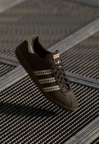 Sneaker in suede marrone con tre strisce marrone chiaro, suola in gomma e parte superiore texturizzata. Include etichetta con marchio sulla linguetta.