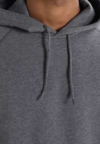 Felpa grigia con una texture morbida, dotata di cappuccio con lacci e maniche raglan. Il tessuto sembra essere una miscela confortevole per un abbigliamento casual.