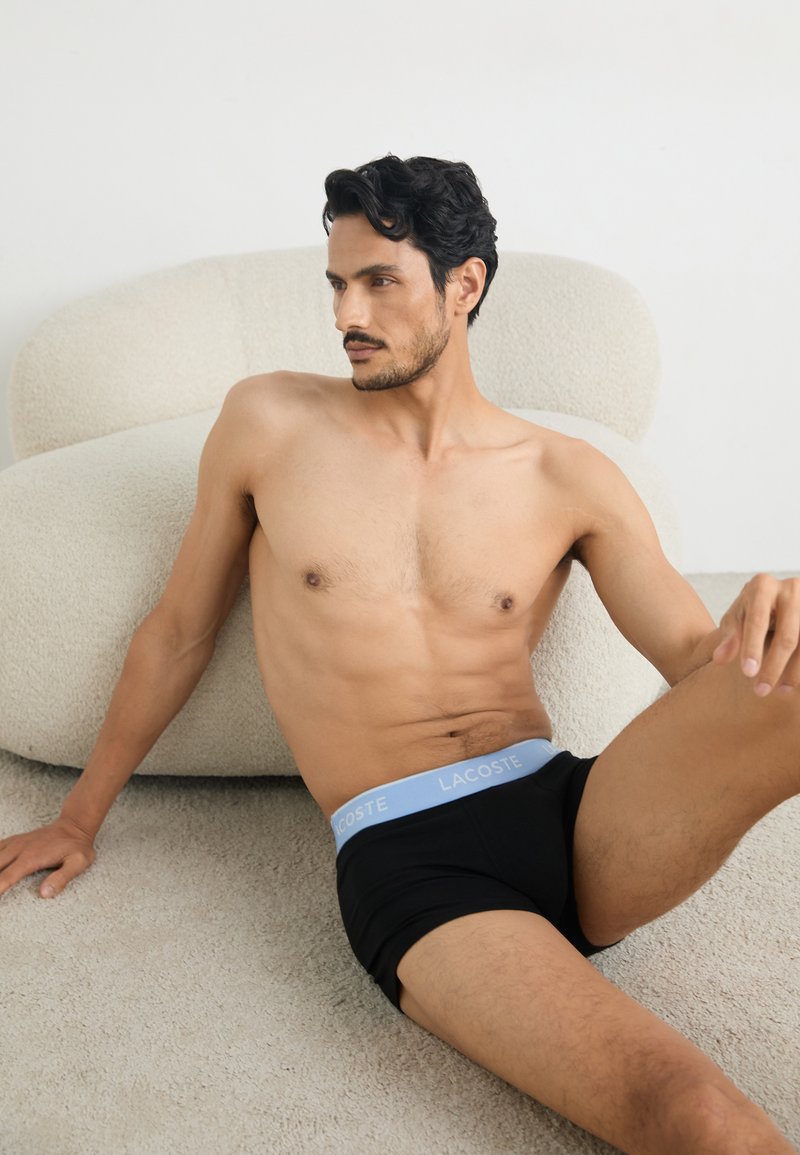 Black boxer briefs with a light blue lace waistband featuring "LACOSTE" logo; model seated on light textured carpet near a soft couch.