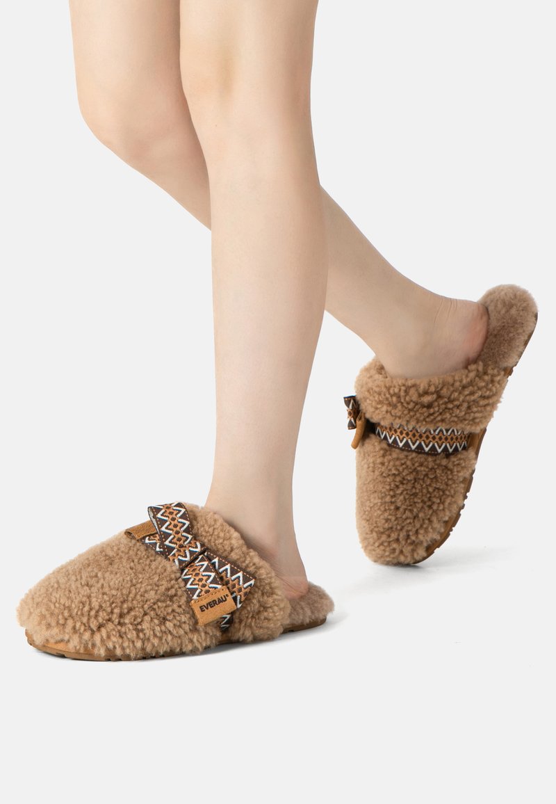 Brown, fluffy slippers with textured wool upper, geometric patterned straps, and a flat sole, designed for comfort and ease of wear.
