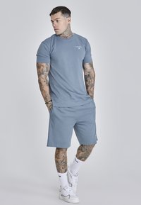 Tricou din bumbac de culoare bleu deschis, cu guler rotund și mâneci scurte, asortat cu pantaloni scurți de aceeași nuanță. Dispune de un logo subtil al brandului și tatuaje. Sneakers albi.