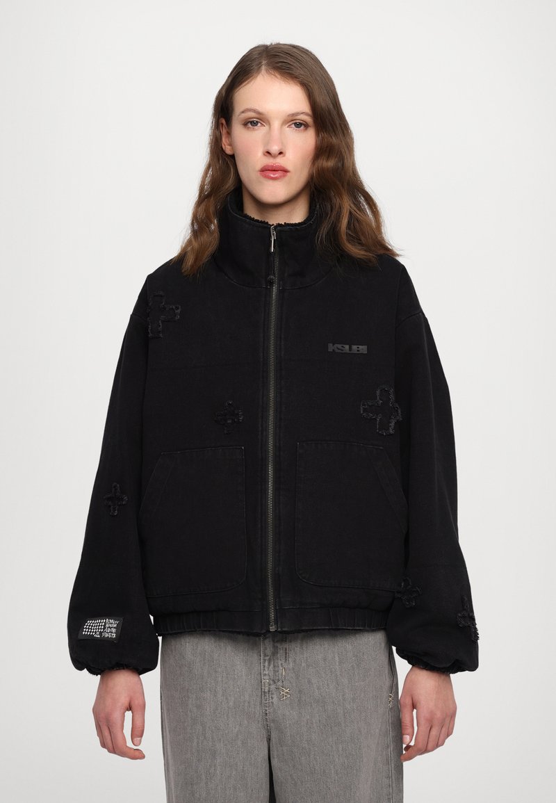 Black denim jacket with a high collar, zip front, and large pockets. Features textured cross patterns and elasticated cuffs.