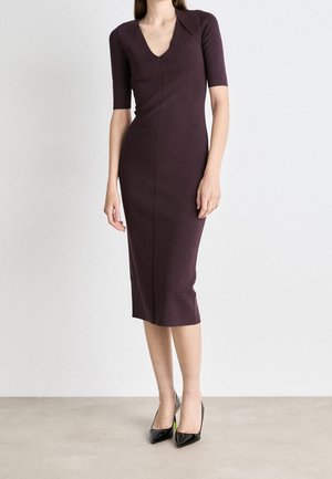 Woman wearing a fitted plum midi dress with elbow-length sleeves and a V-neckline, paired with black pointed-toe heels, standing against a plain wall.