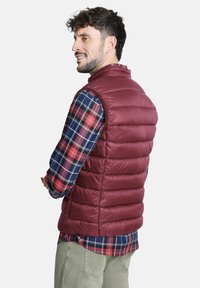 Chaleco acolchado burdeos con costuras horizontales, cuello alto y diseño sin mangas. Llevado sobre una camisa de cuadros de manga larga en azul, rojo y verde.