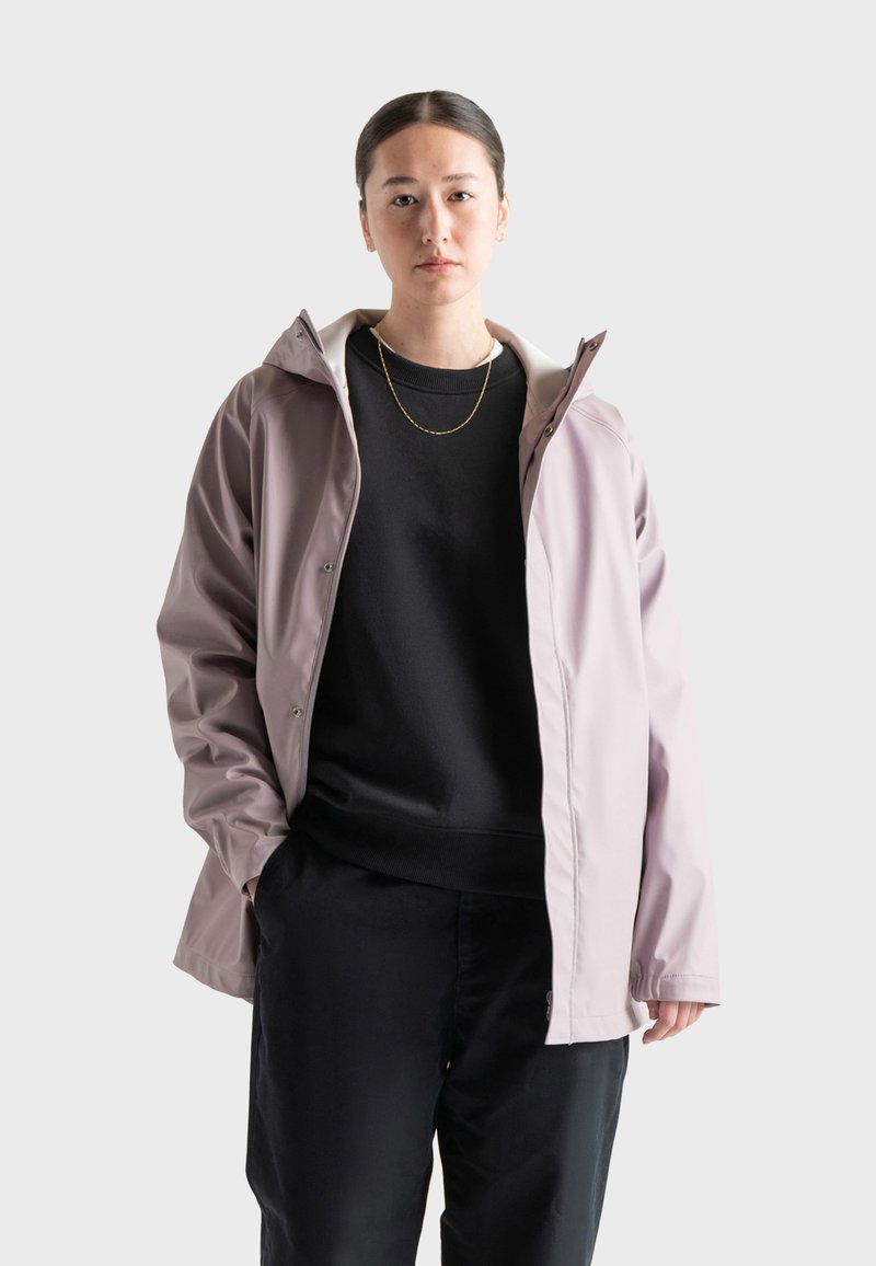 Giacca impermeabile di colore lilla con cappuccio, chiusura frontale con zip e bottoni a pressione, indossata sopra un felpa nera e pantaloni scuri. Texture liscia.