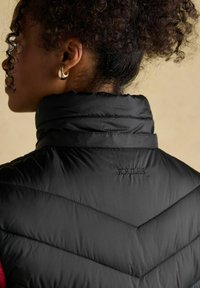 Czarna kamizelka puchowa z wzorem w zygzak, wysoki kołnierz i przeszycia. Na plecach haftowane logo "JOULES".