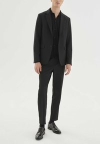 Costume noir sur mesure avec chemise et pantalon assortis. Tissu lisse, coupe ajustée. Chaussures en cuir noir à lacets, détails hardware minimalistes.