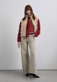 Chaleco de forro polar beige con dos bolsillos, llevado sobre una camisa de manga larga con volantes en rojo intenso. Pantalones anchos de pana color crema y botas marrones con tacón.