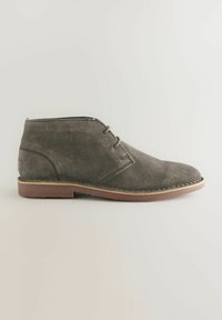 Botín chukka de ante gris con punta redondeada, dos ojetes para los cordones y suela de goma marrón. Presenta costuras visibles a lo largo de la suela.