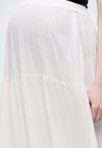 Λευκή υφασμάτινη φούστα με ελαστική μέση που φοριέται από άτομο με το δεξί χέρι ακουμπισμένο δίπλα στο σώμα σε απλό φωτεινό φόντο.