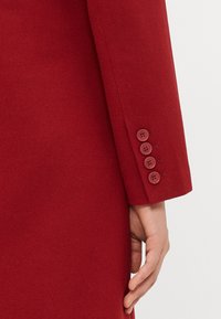 Sleeve of a red coat featuring four matching red buttons, with a hand resting beside it against a white background.
