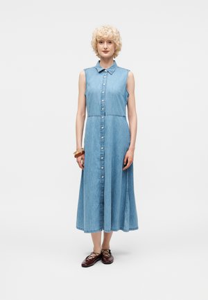 Γυναίκα με κοντά ξανθά σγουρά μαλλιά που φοράει αμάνικο, ανοιχτό τζιν μίντι φόρεμα με κουμπιά και καφέ δερμάτινα παπούτσια με κορδόνια, στέκεται μπροστά σε λευκό φόντο.