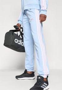 Svetlomodré tepláky s ružovými pruhmi po stranách, v kombinácii s čiernymi športovými topánkami a čiernou taškou na cesty s bielym logom.