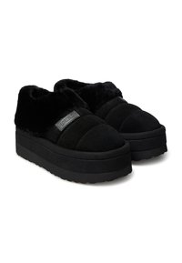 Scarpe slip-on nere con plateau, con una parte superiore morbida e pelosa, in tessuto simile alla pelle scamosciata, e una suola in gomma testurizzata. Presentano un'etichetta di marca prominente.