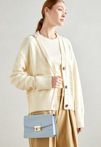 Světle krémový žebrovaný kardigán s knoflíkovým zapínáním, nošený přes béžové tričko. Malá taška přes rameno v nebesky modré barvě obsahuje zlatý řetízkový popruh a sponu.
