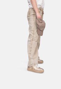 Jeans de denim beige con un corte recto, combinados con zapatillas metálicas doradas y una gorra de cuadros marrón sostenida en la mano.