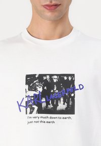 Balta medvilnė marškinėliai su juodai baltu grafiniu spausdiniu, vaizduojančiu minią, ir tekstu „KARL LAGERFELD“ purpurine spalva.