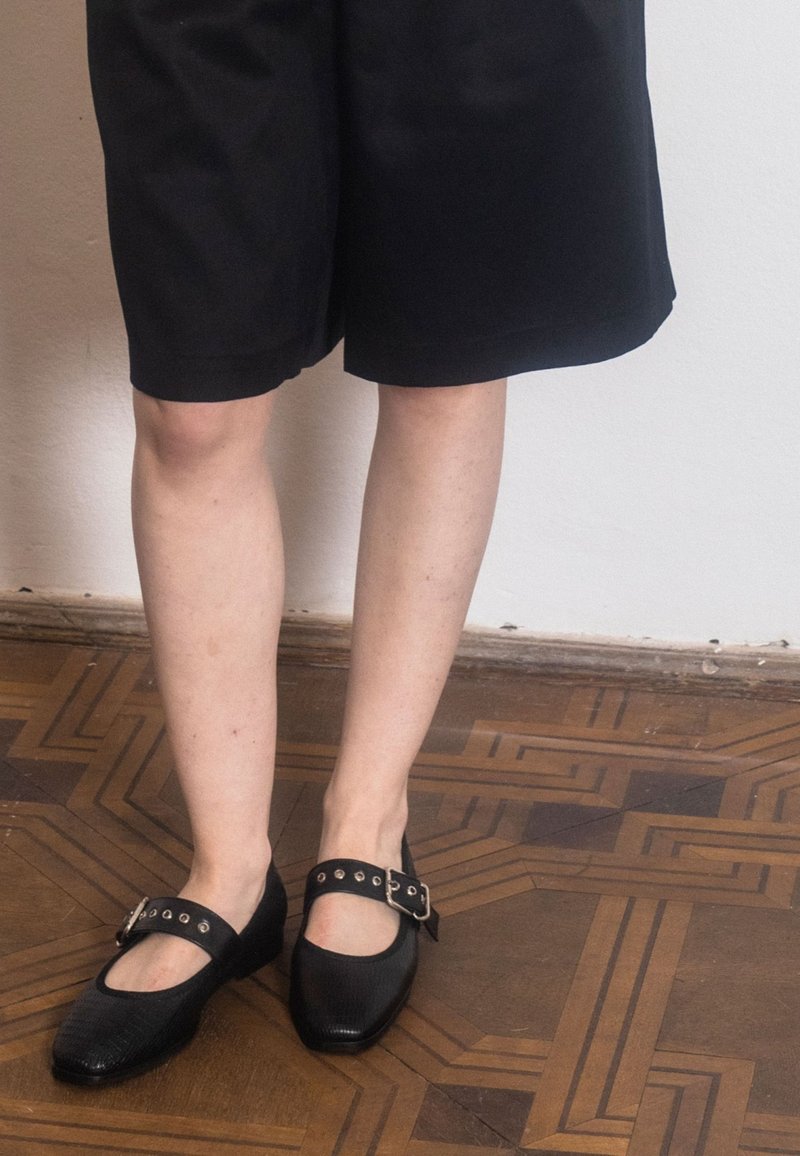 Scarpe piatte nere con punta arrotondata, dotate di una cintura con fibbia e dettagli borchiati; indossate con pantaloni corti neri su un pavimento in legno.
