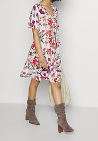 Květinové šaty s krátkými volánovými rukávy, bílé osnovy, růžovými a fialovými květinami, volánkovým výstřihem a odpovídajícím páskem; spárované s volnými šedými botami.