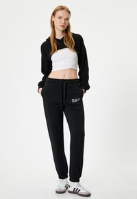Schwarze, kurzhüftige Kapuzenjacke mit langen Ärmeln über einem weißen, eng anliegenden Oberteil, kombiniert mit schwarzen Sweatpants mit Seitentaschen und gesticktem Text.