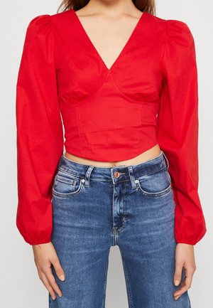 Rotes, kurzes Oberteil mit einem tiefen V-Ausschnitt, gepufften langen Ärmeln und glatter Textur, kombiniert mit blauen, hochsitzenden Jeans mit Knopfverschluss.