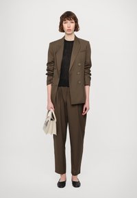 Olijfgroen op maat gemaakt pak met een dubbelgebreide blazer en relaxte broek. Zwarte gebreide top eronder. Model houdt een kleine witte handtas vast.